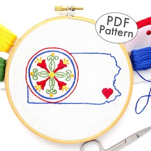 May include: Embroidered Pennsylvania state outline with a red heart in the center, a red, green, and yellow floral design in the upper left corner, and a blue outline. The embroidery is in a wooden hoop with floss spools in red, blue, green, and yellow.
