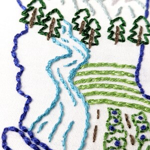 May include: Close-up of an embroidered design featuring a blue river winding through a green landscape with brown trees. The river is surrounded by green hills and a blue border.