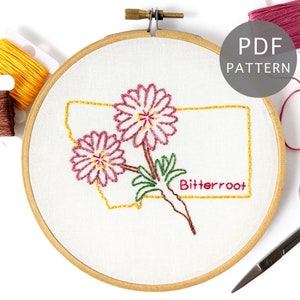 Puede incluir: Arte de bordado en aro con el contorno del estado de Montana con dos flores rosas y la palabra "Bitterroot" en hilo amarillo.
