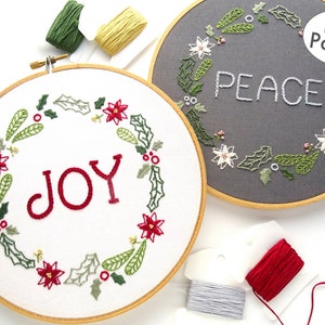 May include: Two embroidery hoops with white fabric and a wooden frame. The hoop on the left has a red and green wreath with the word "JOY" in the center. The hoop on the right has a gray fabric with a white and green wreath and the word "PEACE" in the center.