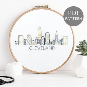 Pode incluir: Um aro de bordado branco com uma cidade de Cleveland, Ohio, bordada em preto e amarelo. O aro está sobre uma superfície branca com uma bola de fio branco e uma tesoura preta.
