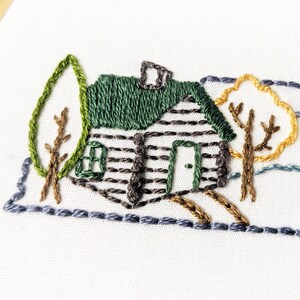 May include: Embroidered design of a cabin with a green roof and brown siding, surrounded by trees with brown branches and green and yellow leaves. The cabin has a small window and a door. The design is stitched on a white fabric.