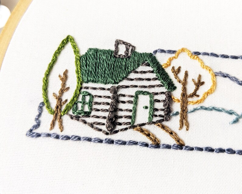 May include: Embroidered design of a cabin with a green roof and brown siding, surrounded by trees with brown branches and green and yellow leaves. The cabin has a small window and a door. The design is stitched on a white fabric.