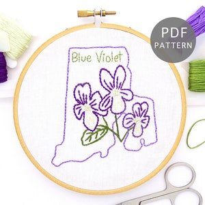 Può includere: Cerchio da ricamo con sfondo in tessuto bianco che presenta il contorno dello stato del Rhode Island con le parole "Blue Violet" e un'immagine ricamata di violette viola.