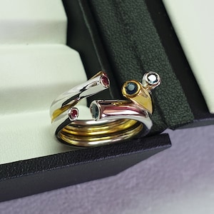 Könnte beinhalten: Ein Satz aus drei Ringen aus Gold und Silber mit bunten Edelsteinen. Die Ringe sind ineinander verschlungen und haben einen blauen, roten und weißen Edelstein.