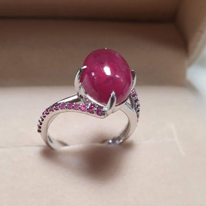 Könnte beinhalten: Ein silberner Ring mit einem großen, ovalen Rubin-Edelstein. Der Ring ist mit kleineren Rubinen um das Band besetzt.