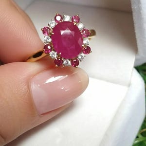 Op de afbeelding: Een gouden ring met een grote, ovaal geslepen robijn. De robijn is omringd door kleinere rode en witte edelstenen, waardoor een bloemachtige halo ontstaat. De ring wordt door een persoon vastgehouden.