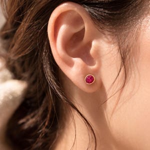 May include: A close-up of a person's ear with a small, round, ruby-colored stud earring set in a gold bezel. The earring is the focal point, with the ear and hair providing a soft background. The earring is a classic jewelry piece.