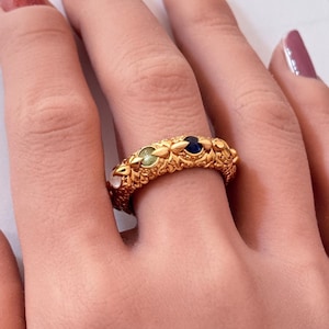 May include: A gold-colored ring with an ornate design featuring small, colorful gemstones. The ring has a textured surface and is worn on a finger. The gemstones are white, green, and blue.