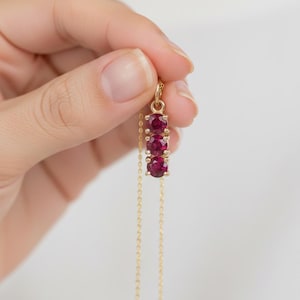 Könnte beinhalten: Eine goldene Halskette mit einem Anhänger aus drei tiefroten, runden Edelsteinen in einer vertikalen Reihe. Die Kette hat eine feine goldene Kette. Der Schmuck wird vor einem weichen, neutralen Hintergrund präsentiert.