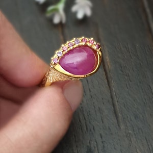 Könnte beinhalten: Ein goldener Ring mit einem großen rosafarbenen Edelstein, der von kleineren rosafarbenen und violetten Edelsteinen umgeben ist.