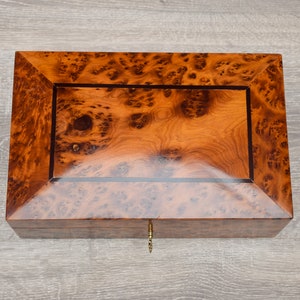 May include: A wooden box with a burl wood finish and a brass keyhole. The box has a dark brown border around the lid.