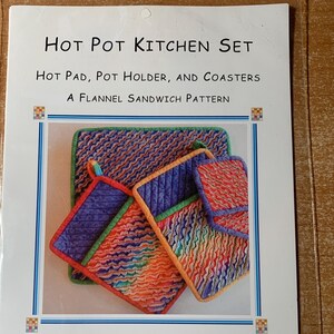May include: A sewing pattern packet titled Hot Pot Kitchen Set for a hot pad, pot holder, and coasters. The cover features a photograph of quilted kitchen items in purple, orange, and blue flannel fabric with a diagonal wavy line pattern.