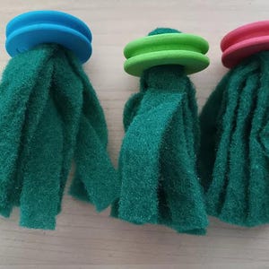 Könnte beinhalten: Drei grüne Schwämme mit farbigen Oberteilen. Die Schwämme haben einen blauen, grünen und rosa Deckel. Die grünen Schwämme bestehen aus einem strukturierten Material. Die Schwämme sind auf einer hellen Holzoberfläche angeordnet.