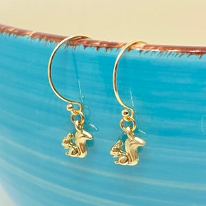 May include: A pair of gold-tone dangle earrings featuring a small squirrel charm. The squirrels are facing to the right and have a bushy tail.