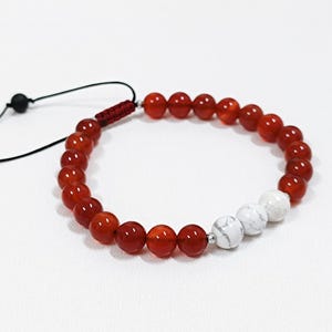 Może przedstawiać: Bransoletka z czerwonych i białych koralików. Czerwone koraliki mają głęboki, bogaty kolor, a białe wyglądają na marmurkowe. Z zapięciem na supeł ślizgowy i czarnymi koralikami na czarnych sznurkach.