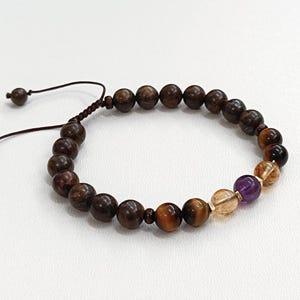 Op de afbeelding: Een kralenarmband met donkerbruine houten kralen en een selectie van gekleurde edelstenen. De armband heeft een schuifknoopsluiting. Edelstenen zijn onder andere tijgeroog, amethist en citrien.