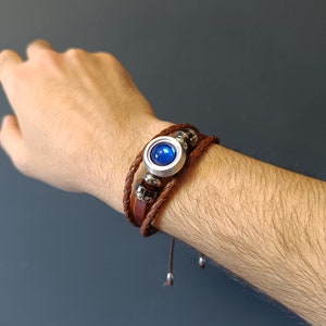 Puede incluir: Un brazalete de cuero marrón con un dije plateado y azul. El dije tiene una piedra preciosa azul en el centro.