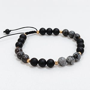 May include: A black beaded bracelet with a sliding knot fastening. The bracelet features matte black and speckled grey beads, interspersed with small gold-coloured beads. The bracelet is displayed on a white surface.