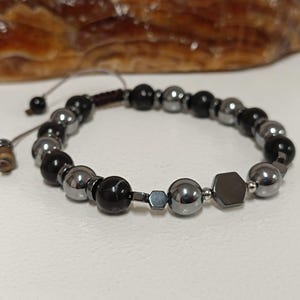 Puede incluir: Una pulsera de cuentas negras, plateadas y gris oscuro. Cuenta con cuentas redondas y hexagonales, ensartadas en un cordón marrón oscuro con extremos ajustables. La pulsera se exhibe sobre una superficie blanca. Las cuentas se miden probablemente en centímetros.