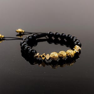 Puede incluir: Una pulsera de cuentas negras con detalles dorados. La pulsera presenta una serie de pequeñas cuentas negras redondas y varias cuentas decorativas doradas. Tiene cordones negros ajustables con extremos dorados.