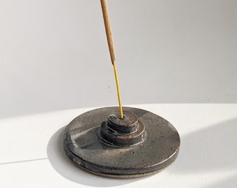 May include: A grey ceramic incense holder with a round base and a raised platform for incense sticks. A brown incense stick is burning and releasing smoke.