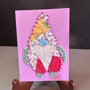 Pode incluir: Um cartão rosa com um gnomo de patchwork colorido feito de retalhos de tecido. O gnomo tem um chapéu vermelho, uma barba branca e um nariz azul. O gnomo é decorado com contas cor-de-rosa.