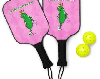 May include: Two pink pickleball paddles with black handles and a cartoon pickle character wearing a crown and holding a purse. The paddles have the text "Pickleball Princess". Two yellow pickleballs are also in the image.
