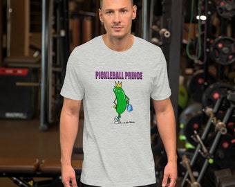 May include: A light gray t-shirt featuring the text "PICKLEBALL PRINCE" above a cartoon pickle character wearing a crown and carrying a briefcase. The pickle is green with yellow accents. The shirt is short-sleeved.