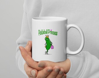 May include: White ceramic coffee mug with a green pickle character wearing a crown and holding a pink purse. The text "Pickleball Princess" is printed on the mug.