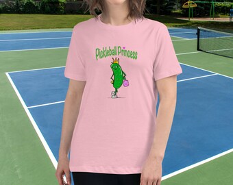 May include: Pink t-shirt with a cartoon pickle wearing a crown and holding a pink bag. The text "Pickleball Princess" is printed on the shirt.