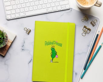 May include: A bright lime green notebook with the words "Pickleball Princess" and a cartoon pickle character. The notebook has a matching elastic band closure and is surrounded by office supplies, including a keyboard and pencils.