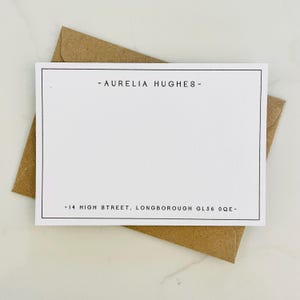 May include: A white card with a black border, featuring the text "- AURELIA HUGHES -" at the top and "-14 HIGH STREET, LONGBOROUGH GL56 0QE-" at the bottom. The card is placed inside a brown envelope.