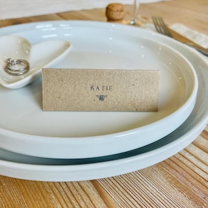 Könnte beinhalten: Ein Gedeck mit weißen Tellern, einer herzförmigen Schale mit Ringen und einer Namenskarte. Die braune Namenskarte trägt den Namen "KATIE" und eine Bienenillustration. Ein Weinglas und ein Korken sind im Hintergrund zu sehen.