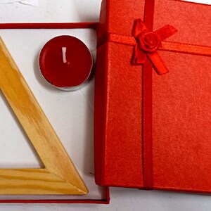 Puede incluir: Un portavelas de madera en forma de triángulo con dos velas de té rojas en una caja de regalo roja con una cinta y un lazo rojos.