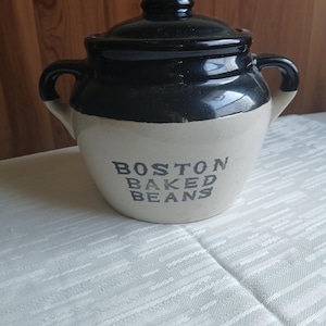 May include: A vintage, two-toned ceramic bean pot with a black lid and rim, and a cream-colored base. The words "BOSTON BAKED BEANS" are printed in dark gray on the front. The pot has two handles and sits on a white tablecloth.