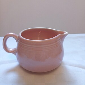 May include: A light pink ceramic creamer featuring a rounded body, a small pouring spout, and a single curved handle. The exterior of the vessel is decorated with a series of horizontal embossed rings encircling the upper portion of the piece.