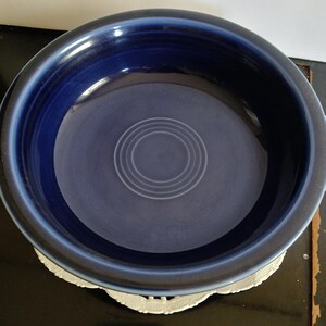 May include: A deep blue ceramic bowl with a circular design in the center. The bowl has a wide rim and a smooth, glossy finish. The bowl is sitting on a white, lacy doily.
