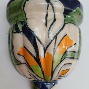 May include: Hand-painted ceramic wall pocket in a bulbous shape. The design features a large white flower with orange and green accents, outlined in black. The rim and base are dark blue, and the leaves are green.