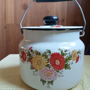 May include: White enamelware pot with a black handle and lid. The pot features a floral design with red, yellow, and white flowers. A metal handle arches over the top. The pot sits on a yellow and white checkered tablecloth.