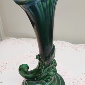 May include: A tall, green ceramic vase with a flared top and a curved, decorative base. The vase has a glossy finish and a flowing, organic design. The color transitions from a deep green to a lighter, blue-green at the top.