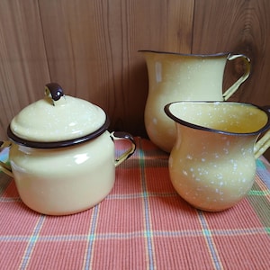 May include: A set of three vintage enamelware kitchen items: a lidded sugar bowl, a pitcher, and a small creamer. The items are yellow with brown trim and white speckles. The set is displayed on a red and green plaid tablecloth.
