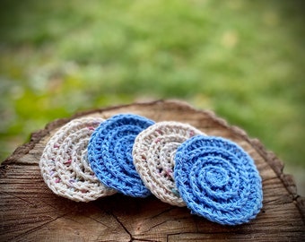 May include: Four crocheted coasters, two in a light beige with white and pink flecks and two in a light blue. The coasters are arranged in a semi-circle on a wooden surface.