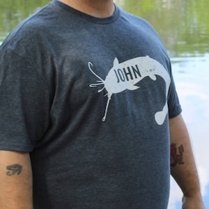 May include: A man wearing a tie-dye bandana and a dark grey t-shirt with a white catfish graphic and the word "JOHN" printed on it. He is standing in front of a pond with a fountain in the background.