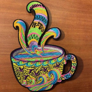 May include: A colorful embroidered patch of a steaming cup of hot tea. The cup is yellow with blue, green, and pink swirls. The steam is multi-colored and has the words "Hot Tea" embroidered in green.