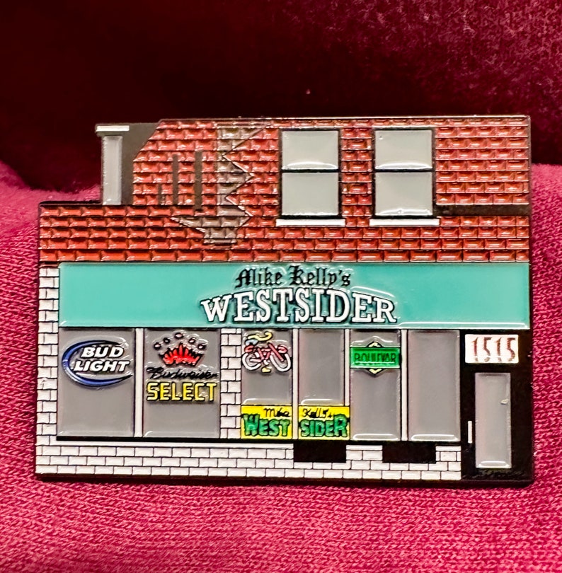 Puede incluir: Un pin de metal que representa una tienda con un toldo verde que dice "Mike Kelly's Westside". La tienda tiene una fachada de ladrillo y ventanas con varios logotipos, incluidos Bud Light, Budweiser Select y una bicicleta. La puerta tiene el n&uacute;mero "1515" en ella.