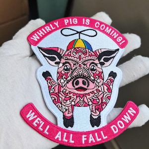 May include: Embroidered patch featuring a pink pig with intricate patterns and a colorful hat. The patch has the text "WHIRLY PIG IS COMING!" above the pig and "WE'LL ALL FALL DOWN" below, all in pink lettering.