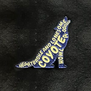 May include: A blue glitter enamel pin in the shape of a howling coyote. The pin features the words "Heartbroke and Lonesome Coyote Howl A-00000" in yellow lettering, creating a unique design. The pin is set against a dark background.