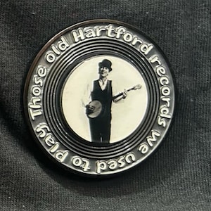 May include: A silver-toned round pin with the text "Those old Hartford records we used to play" encircling a black and white image of a man playing a banjo. The man is wearing a vest and hat.