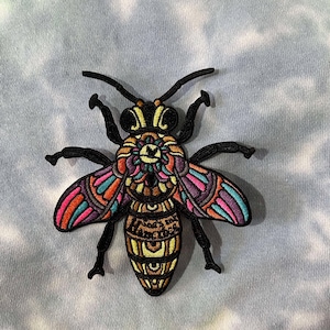 May include: A colorful embroidered bee patch with intricate details. The bee's wings are a vibrant mix of pink, orange, blue, and purple. The bee's body is a golden yellow with black outlines and a mandala-like design. The text "Love is the answer" is embroidered on the bee's abdomen.